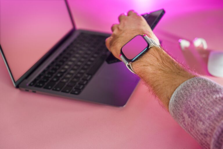 Close-up of a hand using a smartphone, smartwatch, and laptop on a pink desk.